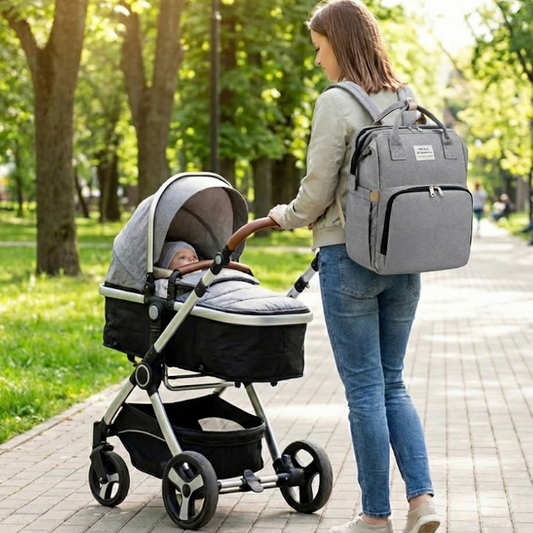 Woman pushing a stroller with a baby in a park, carrying a matching gray backpack.